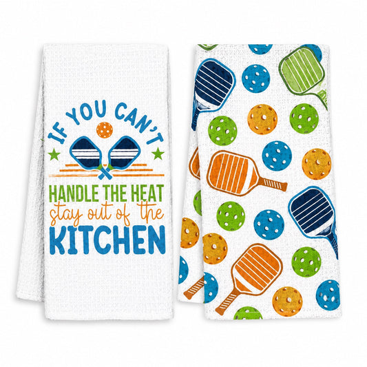 Two white kitchen towels shown on a plain white background. The left towel has a printed slogan reading 'IF YOU CAN'T HANDLE THE HEAT stay out of the KITCHEN' with crossed paddle balls and blue stars.