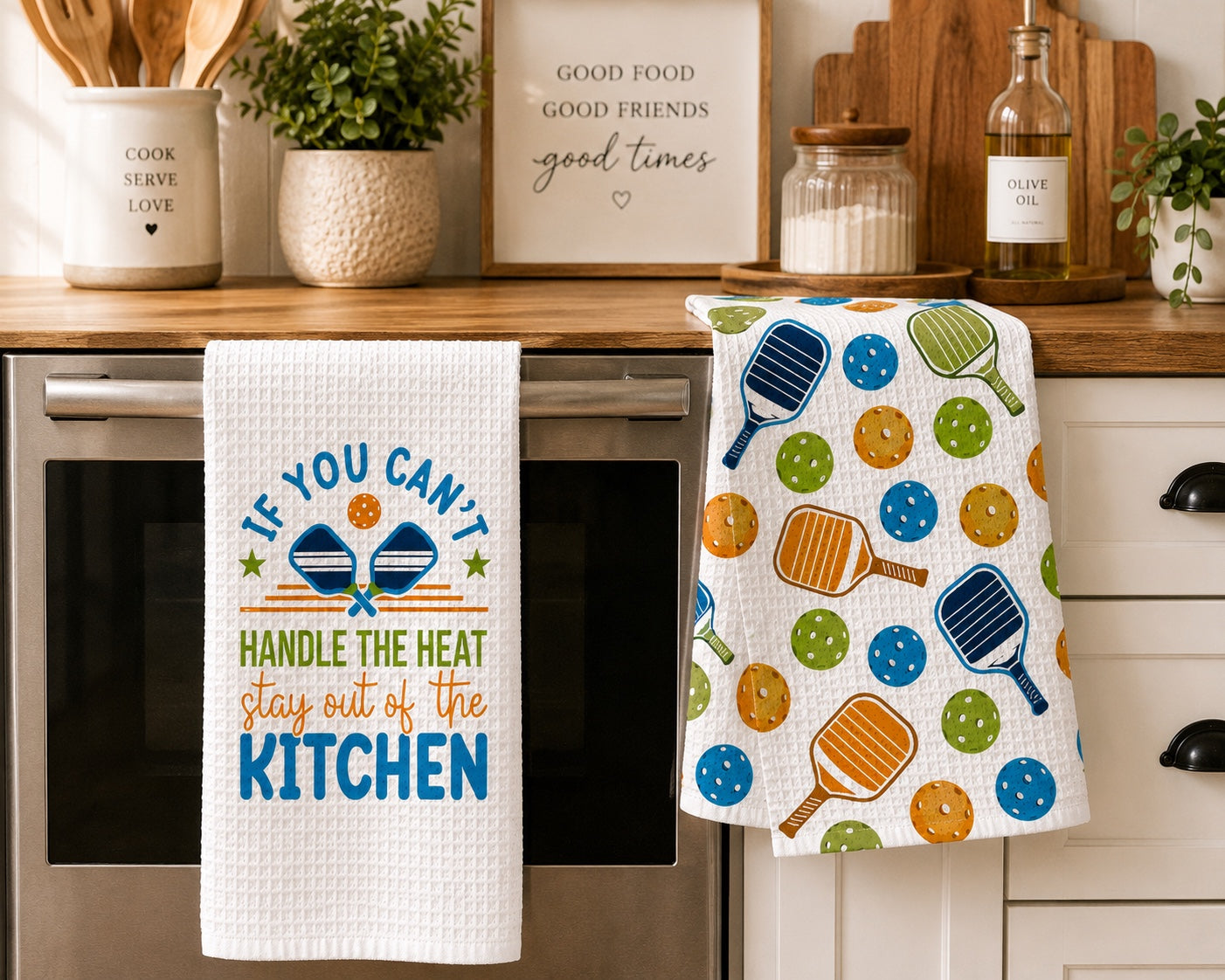 Two kitchen towels hanging on an oven handle in a real kitchen setting. The left towel is white waffle-textured with a blue and orange text design reading 'IF YOU CAN'T HANDLE THE HEAT, stay out of the KITCHEN.