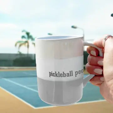 Person holding a mug with 'pickleball peo' text by a tennis court