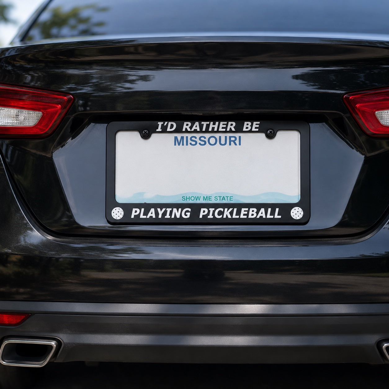 A black license plate frame mounted on the rear of a black car, featuring the text 'I'D RATHER BE MISSOURI!' at the top and 'PLAYING PICKLEBALL' at the bottom with 'SHOW ME STATE' in blue across the middle.