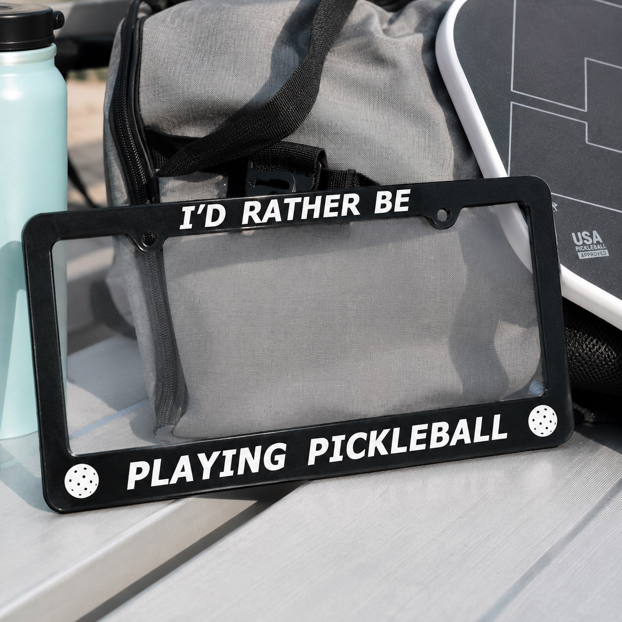 A black license plate frame with white text reading 'I'D RATHER BE PLAYING PICKLEBALL' and small white pickleball dot graphics in the corners, shown outdoors on a table with a gray bag and other items in the background.