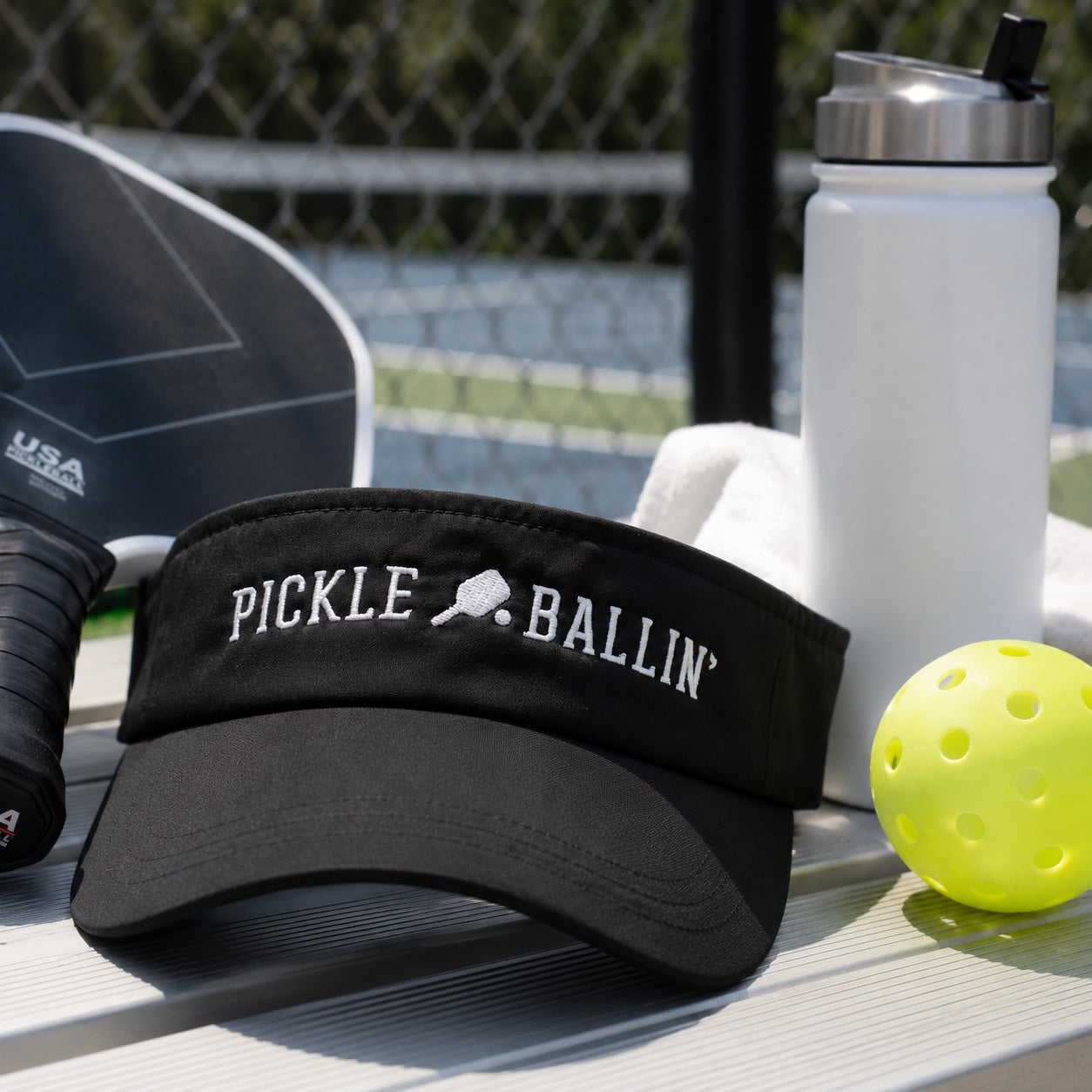 A black visor with 'PICKLE BALLIN'' embroidered in white sits on a picnic table outdoors, surrounded by pickleball gear including a paddle, a white water bottle, and a yellow pickleball.