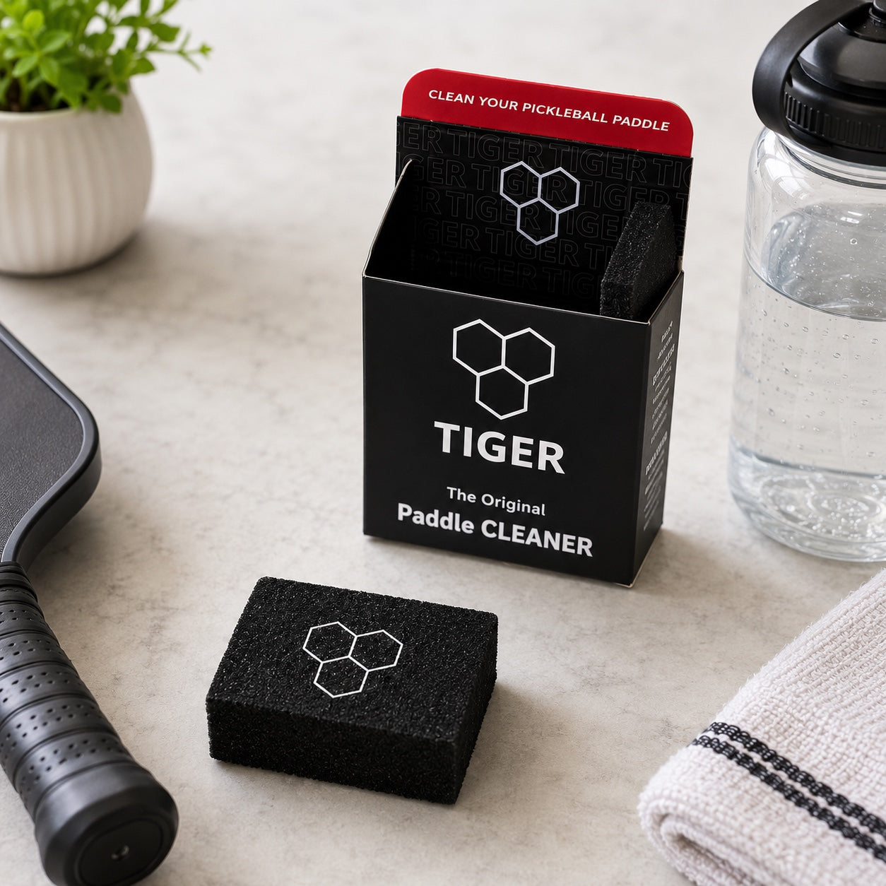 A black sponge pickleball paddle cleaner box labeled 'TIGER' stands open on a light stone surface, with a matching black sponge in front and a pickleball paddle and water bottle nearby as contextual props.