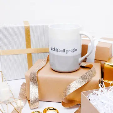 White mug with 'Pickleball people' text on a gift box with gold ribbon, surrounded by wrapped gifts.