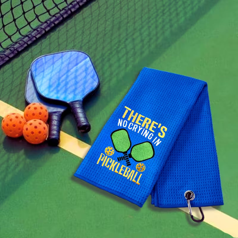 A blue pickleball-themed towel with embroidered text and graphics is displayed on a green pickleball court next to two paddle shovels and orange balls.