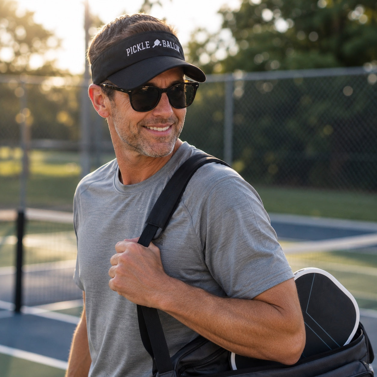 A man on a tennis court wearing a black visor with 'PICKLE BALL' printed in white, black sunglasses, a gray t-shirt, and carrying a black and white sports bag over his shoulder.