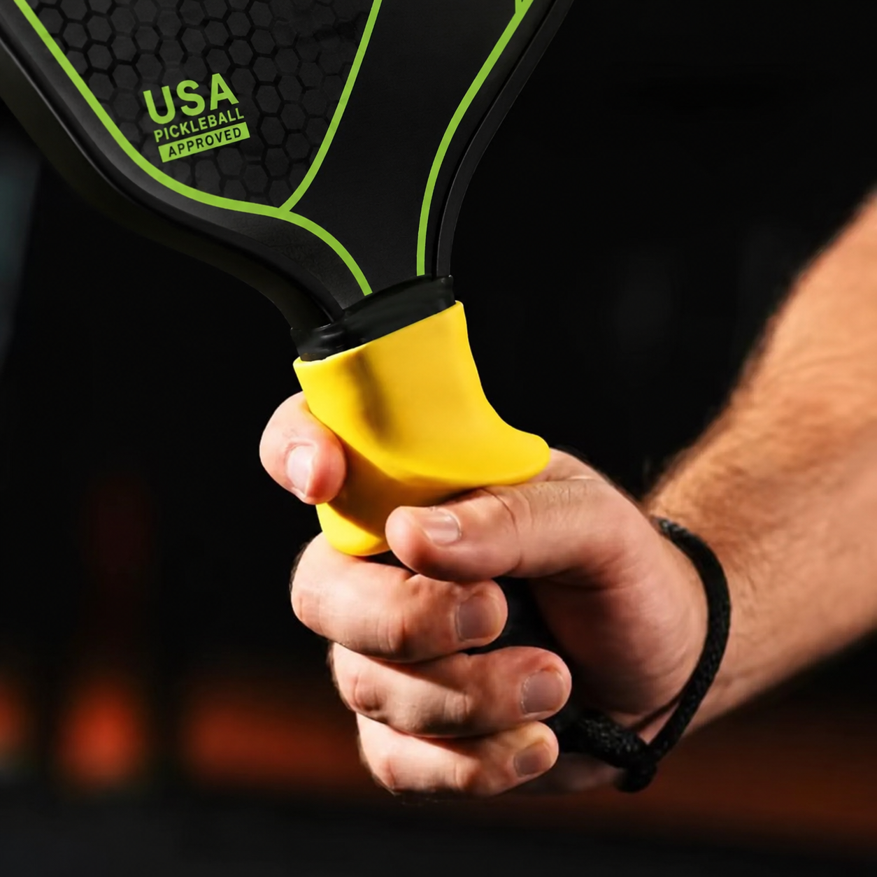A close-up of a person's hand gripping the yellow and black handle of a pickleball paddle, with the paddle blade showing a black honeycomb pattern and green trim. The paddle has 'USA PICKLEBALL APPROVAL' printed on it.