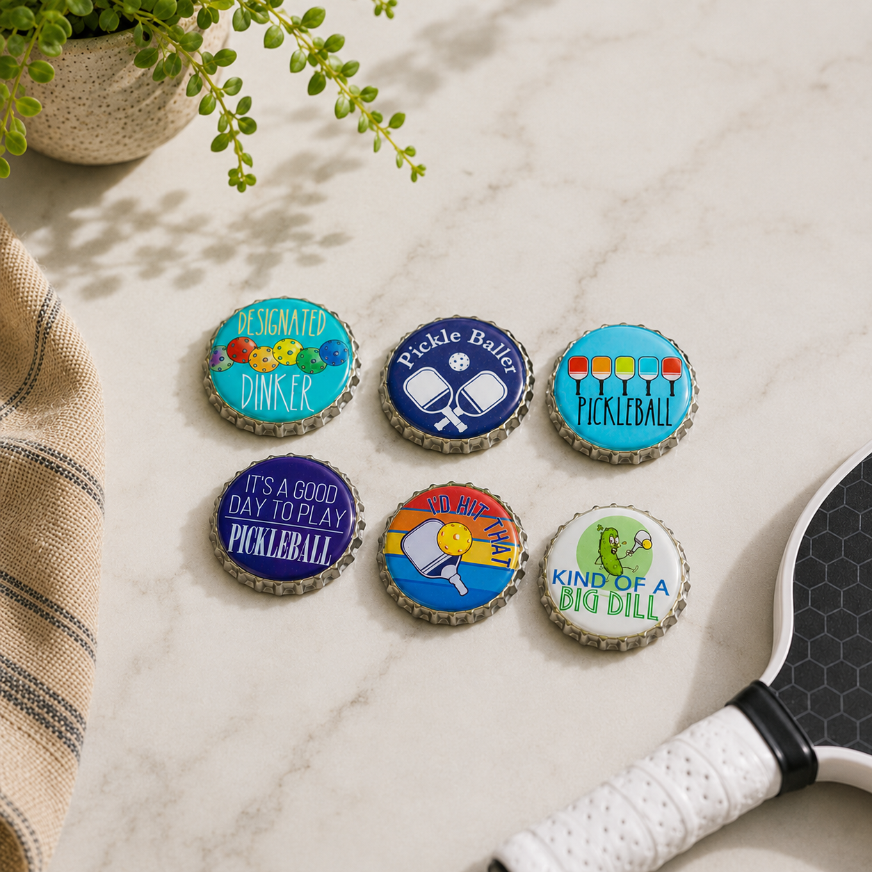 Six pickleball-themed bottle cap coasters arranged on a light marble surface, with part of a paddle and a plant visible in a styled setting.