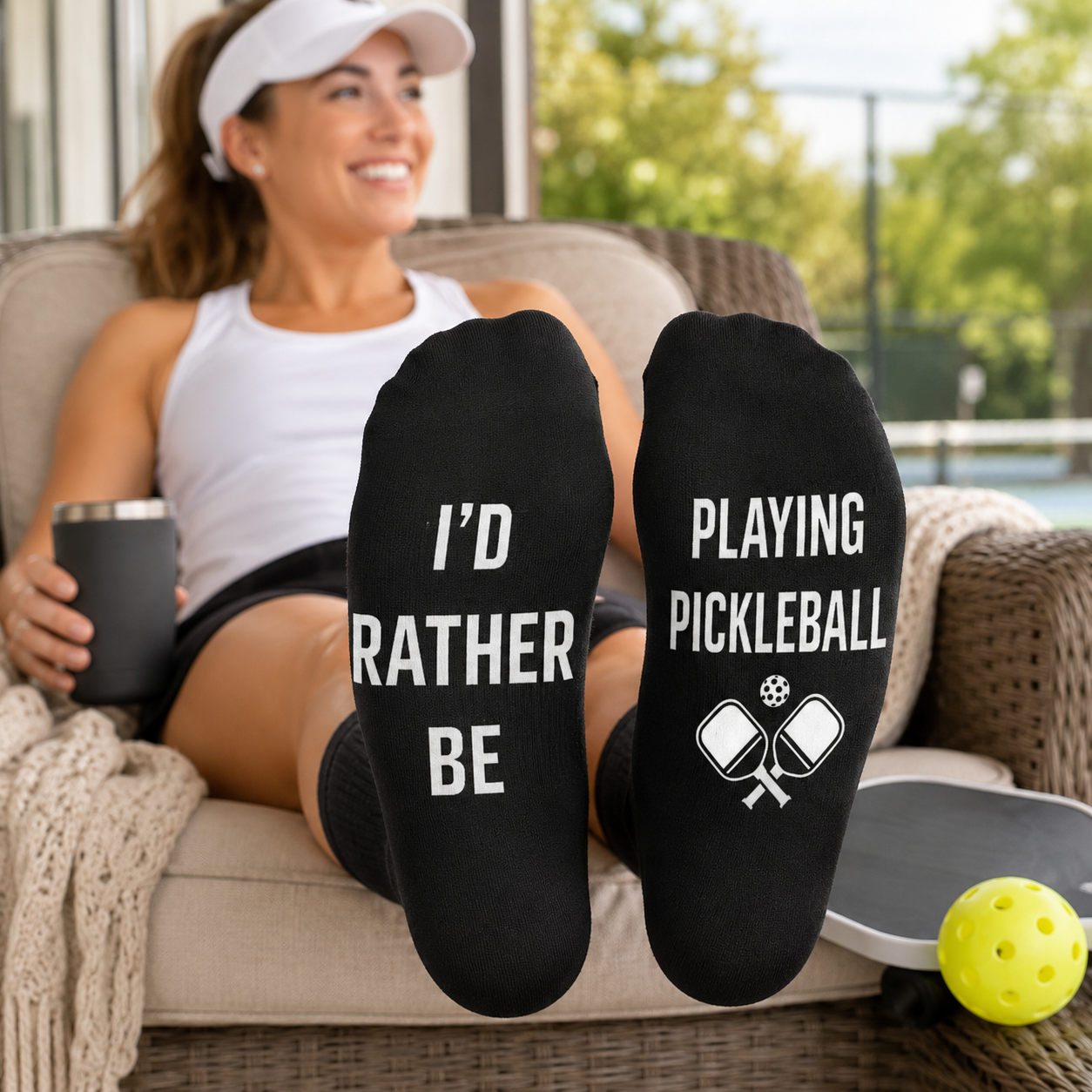 A woman relaxes on a patio chair with her feet up, wearing black socks printed with white text that reads 'I'D RATHER BE PLAYING PICKLEBALL' along with crossed pickleball rackets. A yellow pickleball and a metal tumbler are visible in the scene.