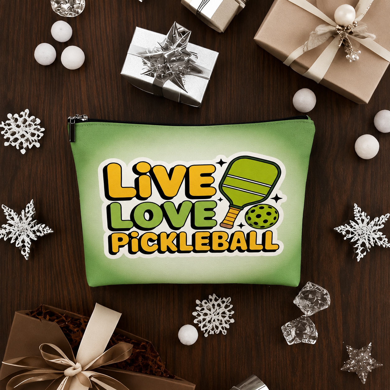 A green zippered pouch with a 'Live Love Pickleball' graphic, styled in a flat-lay on a dark wood background surrounded by holiday gift boxes, snowflake ornaments, and white decorative balls.