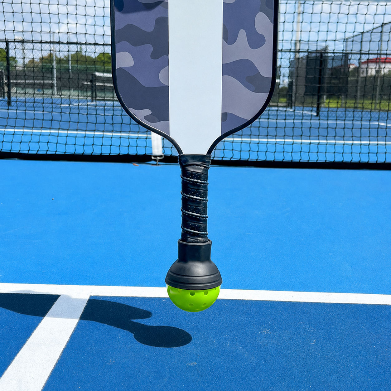 A pickleball paddle with a gray and white camouflage pattern and a black wrapped handle is shown holding a bright yellow pickleball on a blue outdoor court with a net in the background.