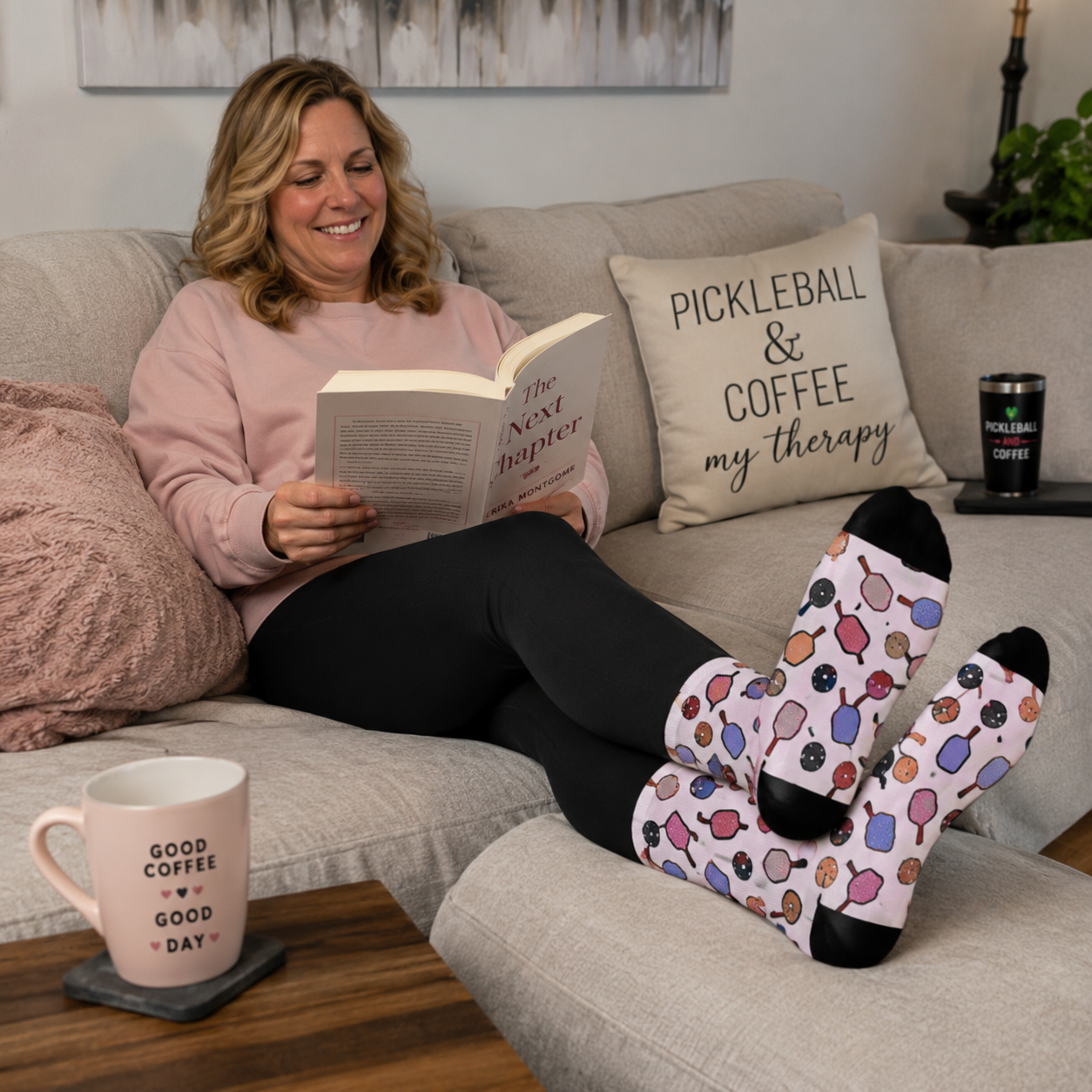 A woman relaxes on a light gray sofa reading a book, wearing pink socks with a colorful donut pattern. A pink mug on a wooden table in the foreground reads 'GOOD COFFEE', and a decorative pillow on the couch says 'PICKLEBALL & COFFEE my therapy'.