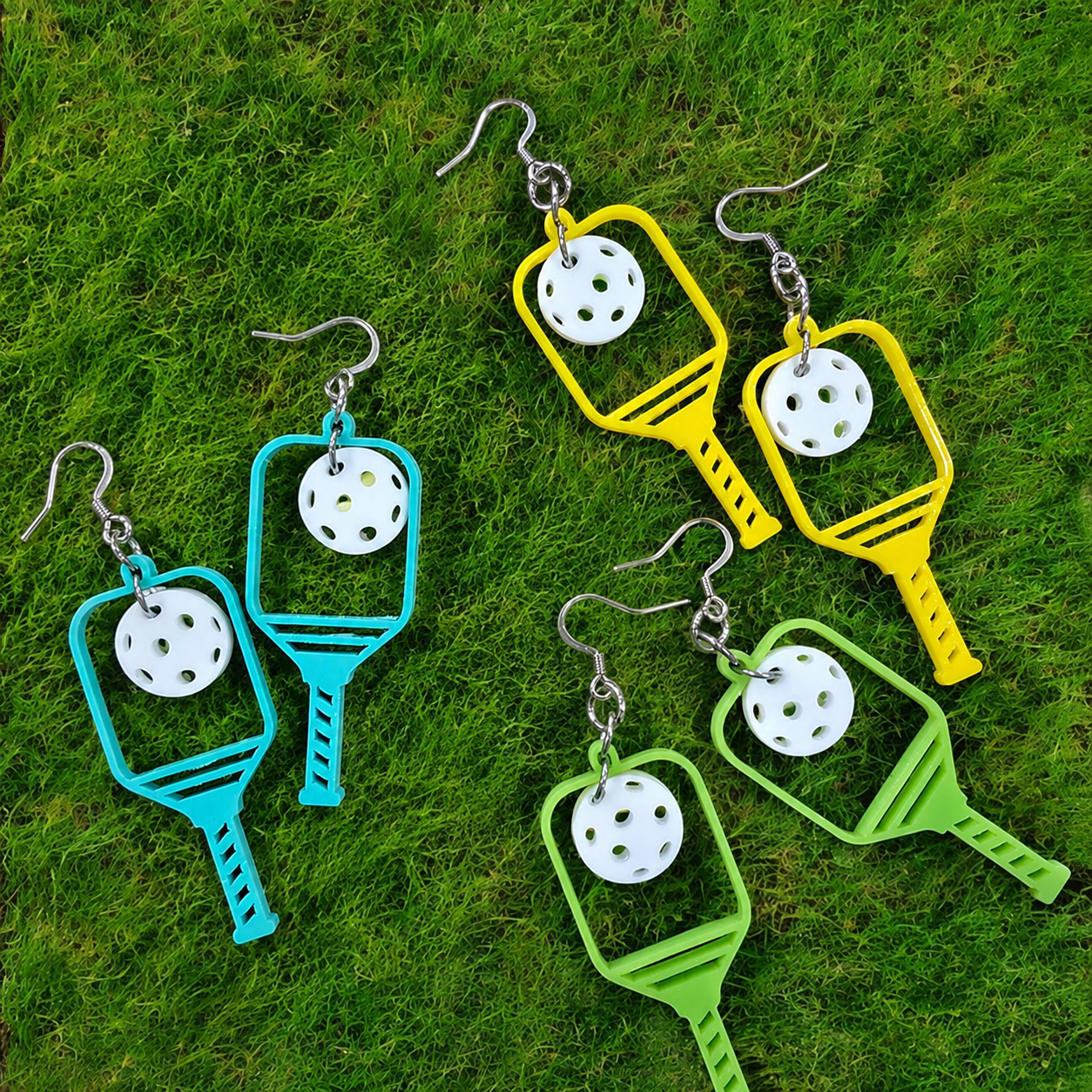 Four pairs of dangling earrings laid on green grass. Each earring has a silver fishhook ear wire and a tennis racket-shaped charm in bright turquoise, yellow, and green, with a white perforated disc hanging in the center.