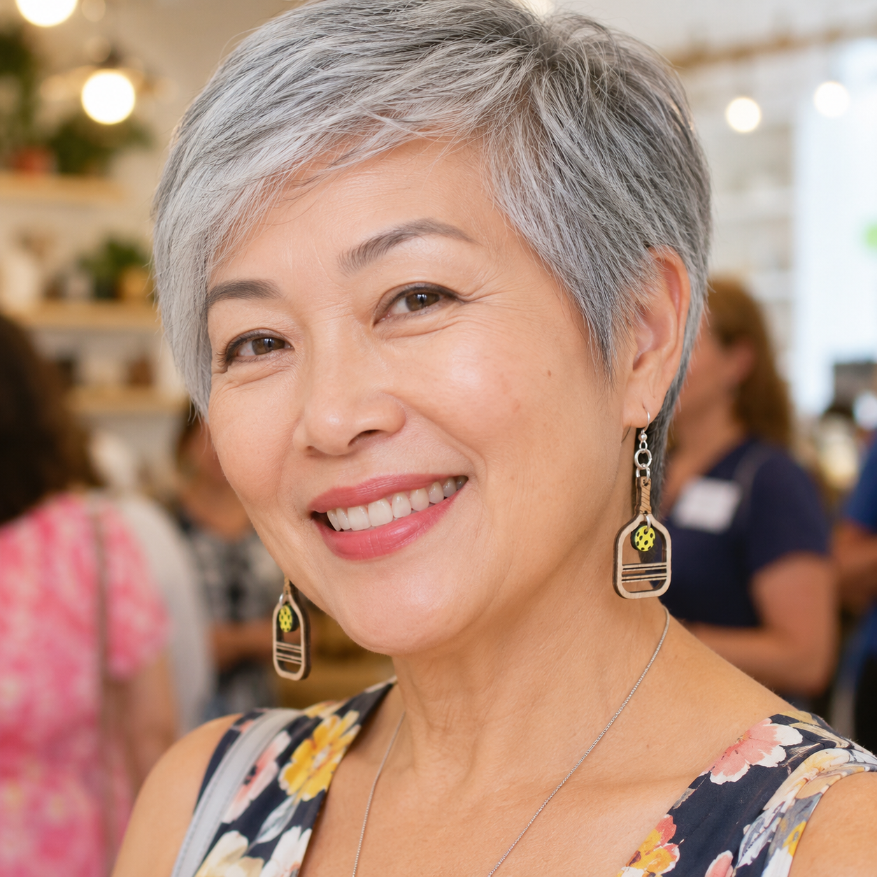 A smiling woman with short gray hair wearing long geometric dangling earrings with gold and green details, photographed in a softly blurred indoor setting.