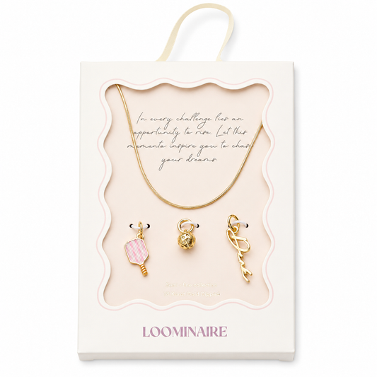 A boxed jewelry set from Loominaire featuring a gold necklace and three gold charms with pink and gold details, presented in a cream-colored display box with a hanging loop.