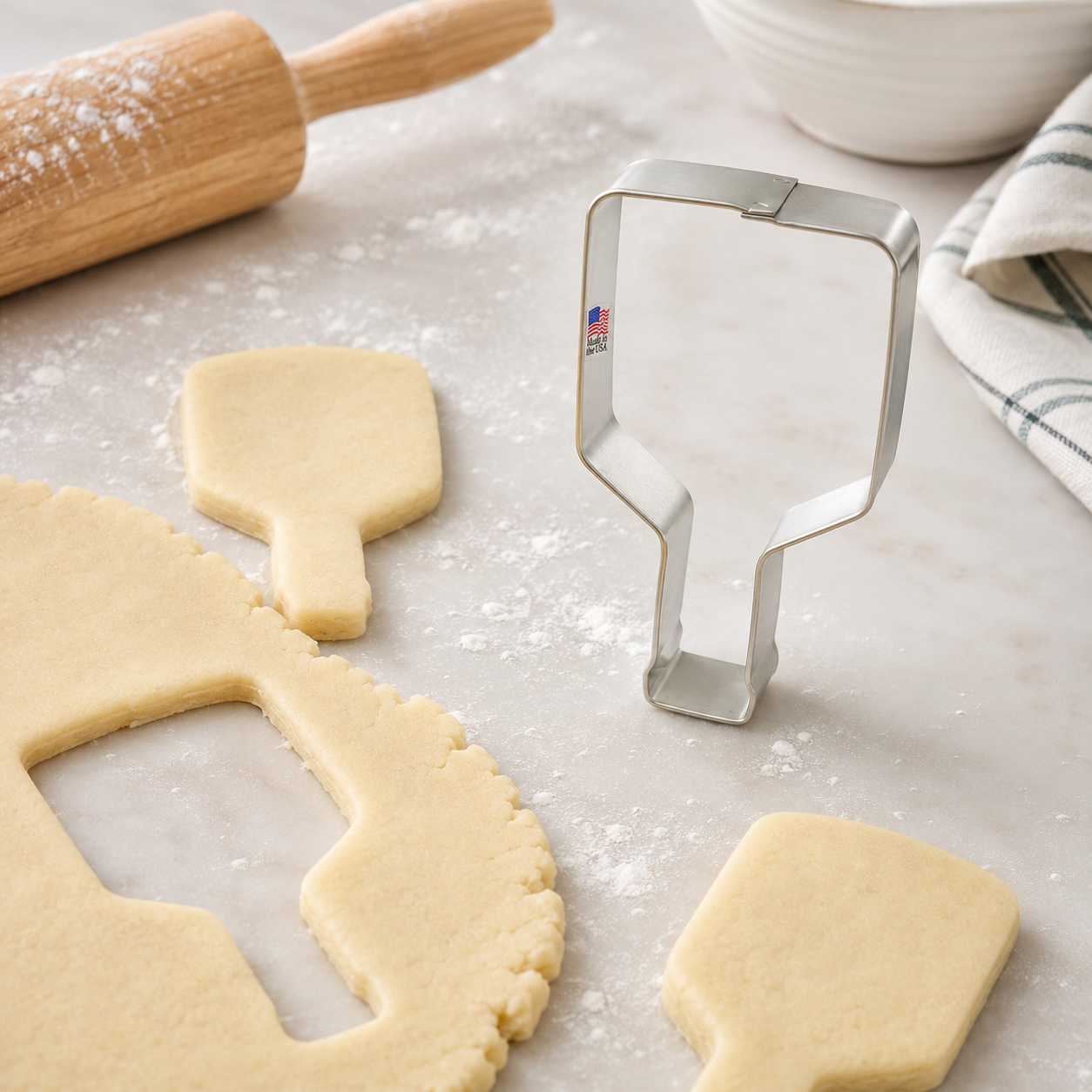 A stainless steel bottle-shaped cookie cutter stands on a light marble surface beside rolled-out dough and cookie cutout pieces, with a wooden rolling pin and white bowl in the background.