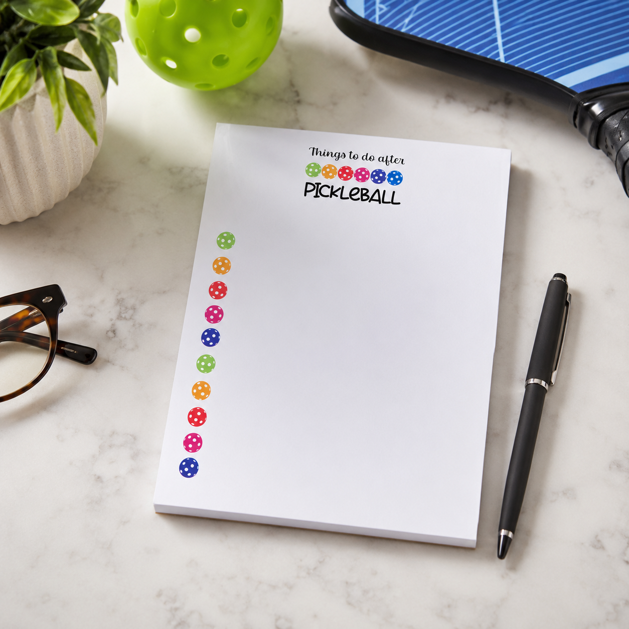 A white notepad with the words 'Things to do after PICKLEBALL' and colorful pickleball-themed dots printed on it, styled on a marble surface with a black pen, plant, and other desk props nearby.