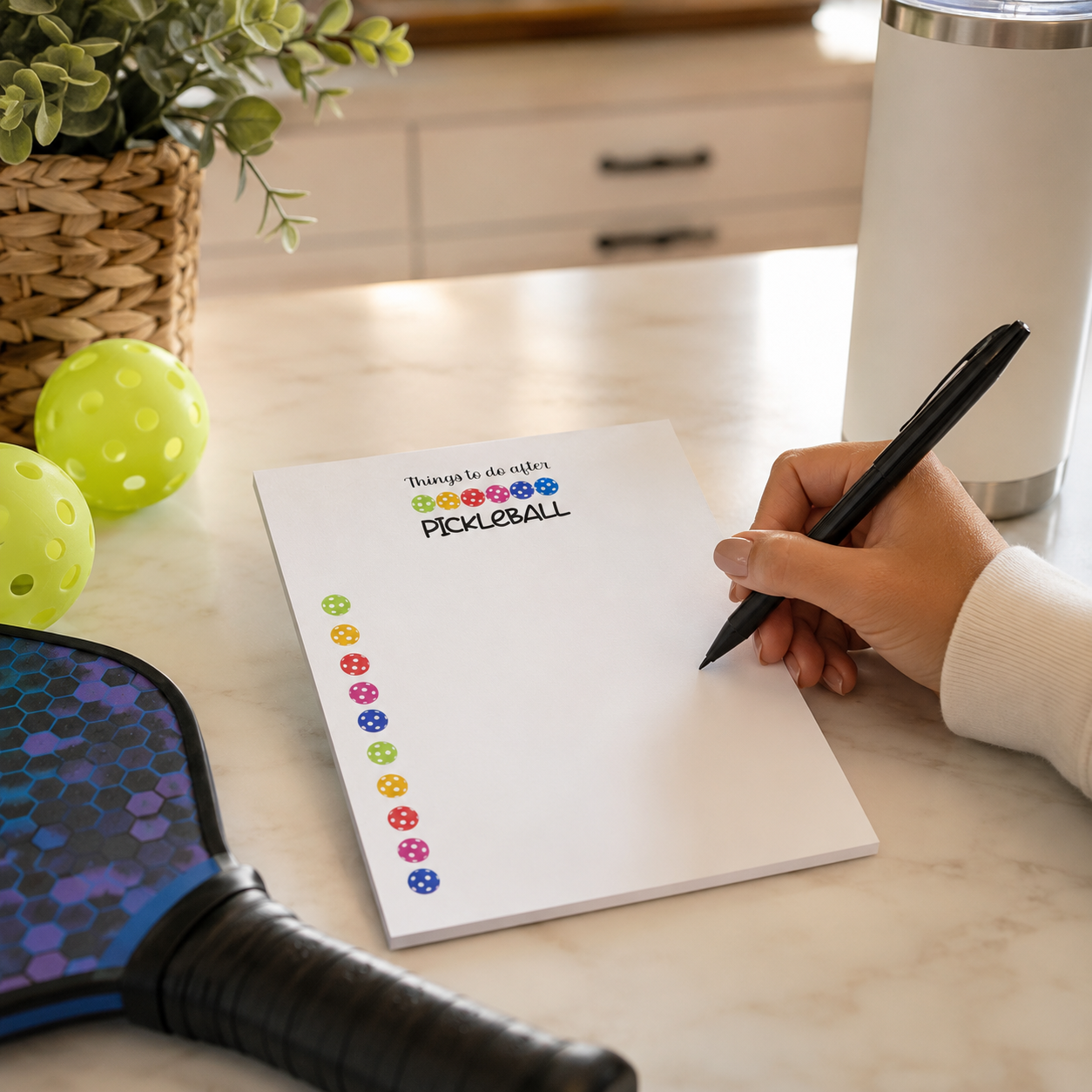 A hand holding a black pen writes on a white notepad that says 'Buena tarde! PICKLEBALL' next to colorful round stickers. A pickleball paddle and two lime balls sit on a marble countertop in a kitchen setting.