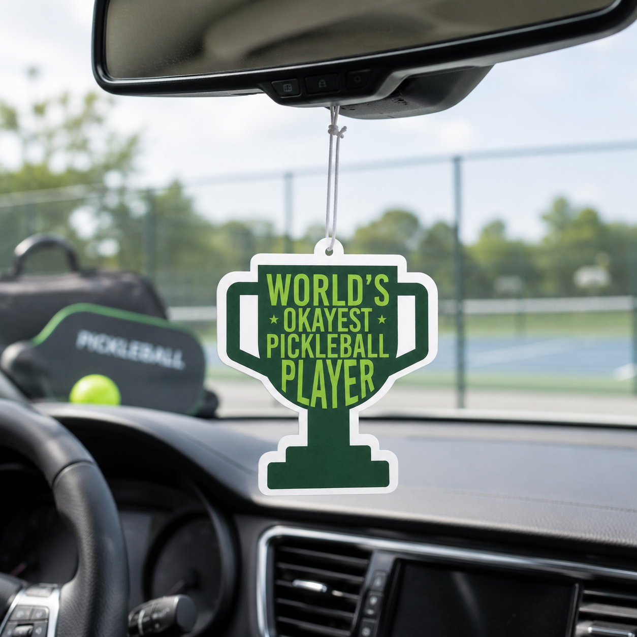 A trophy-shaped car air freshener hanging from the rearview mirror in a car. The air freshener is dark green with a white border and lime green text that reads 'WORLD'S OKAYEST PICKLEBALL PLAYER'.