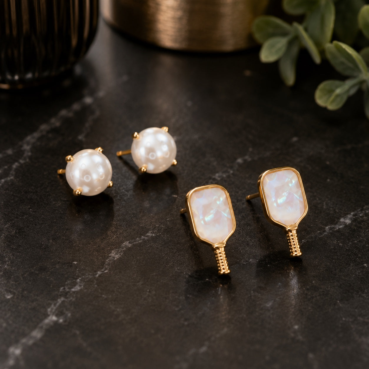 Three gold-toned stud earrings displayed on a dark marble surface: two round pearl-like studs with gold prongs and one rectangular iridescent white stud with a gold textured post. Decorative plants and vessels are blurred in the background.