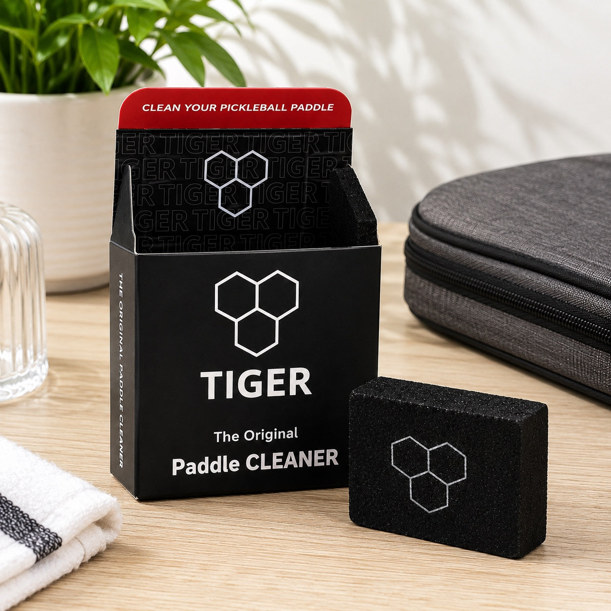 Tiger Paddle Cleaner set on a light wood tabletop, featuring a black and red product box, a black foam cleaning block, a gray zippered case, a folded white towel, and a glass jar in a styled indoor setting.