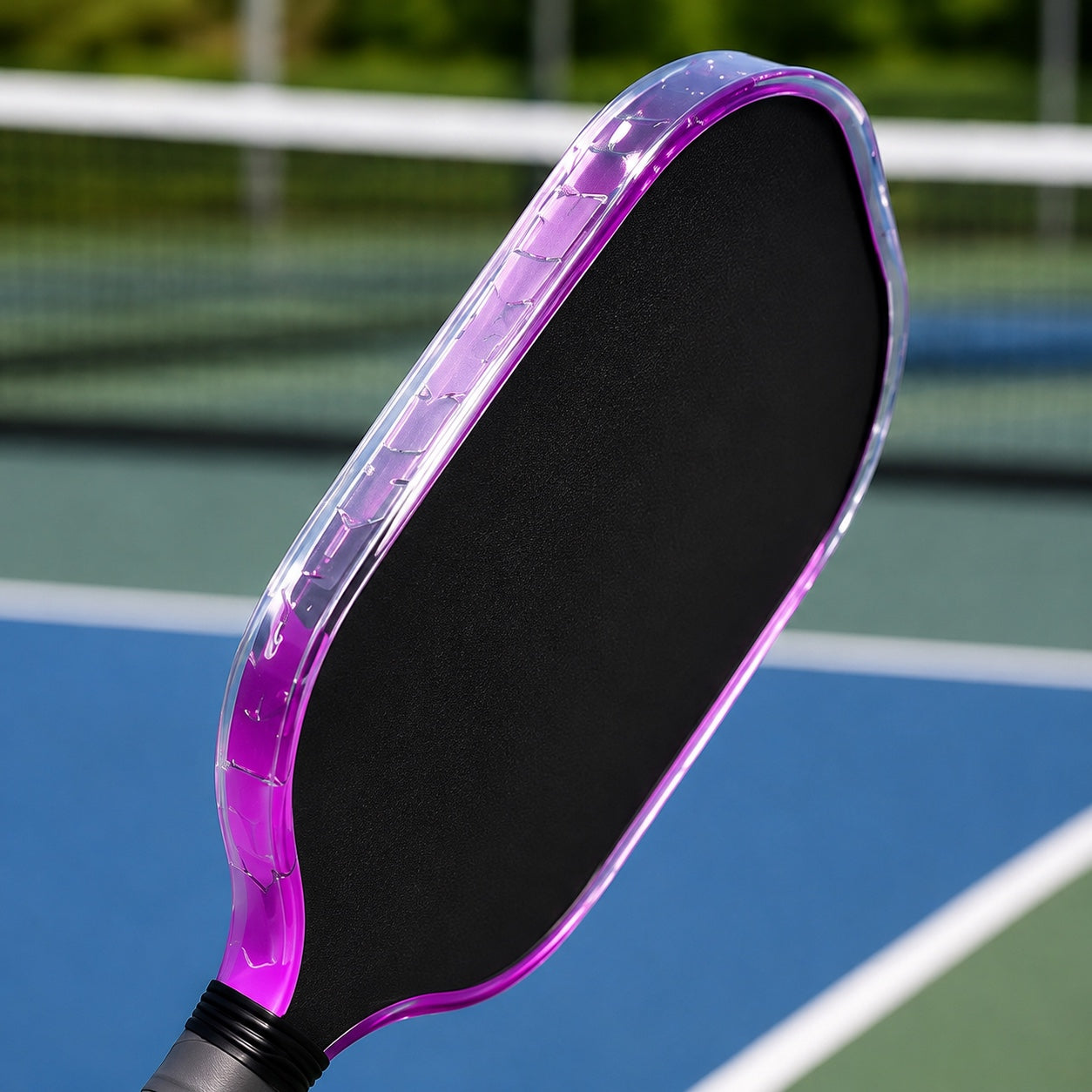 A close-up of a pickleball paddle with a black textured paddle face and a translucent purple edge, shown on an outdoor pickleball court with a blue court surface and green netting in the background.