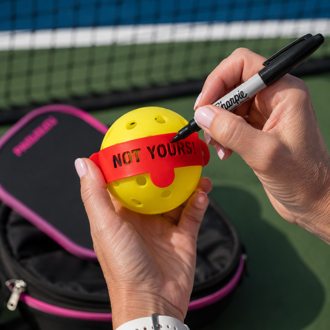 A yellow pickleball with a red custom band reading 'NOT YOURS!' being written on with a black marker, held by two hands on a tennis court with a pink and black bag in the background.
