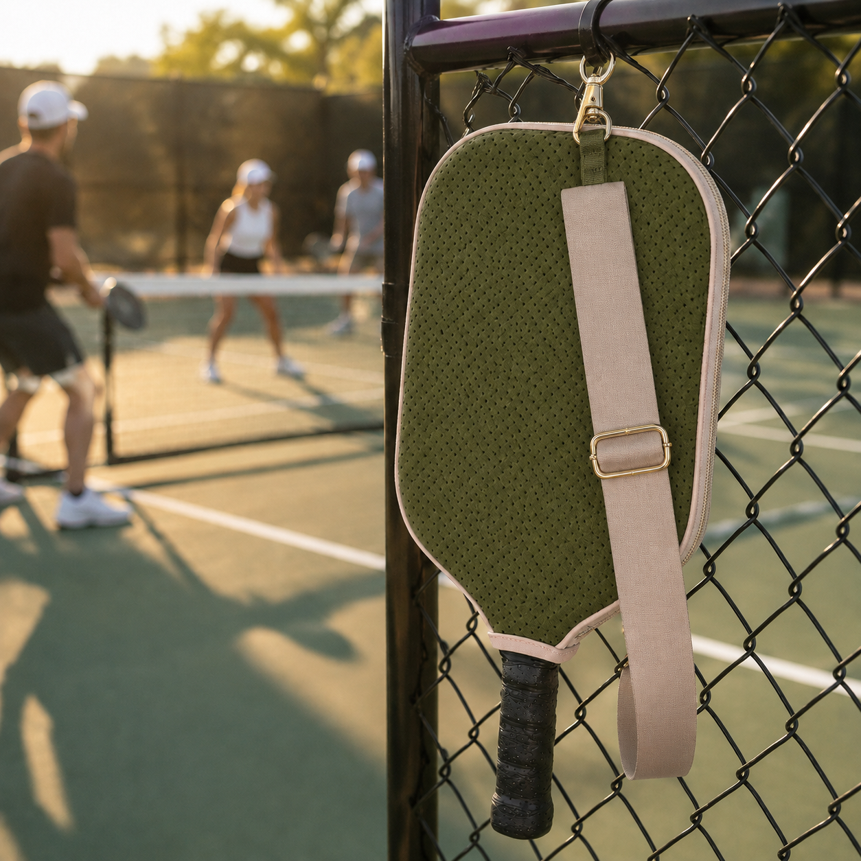 Olive green pickleball paddle cover hanging from a court fence by its built-in gold clip