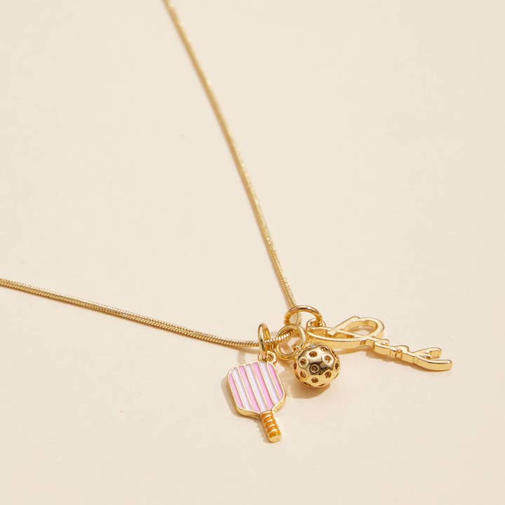 A gold-tone necklace with three charms: a pink and white striped paddle-shaped charm, a gold ball charm with black dots, and a small gold key charm, displayed on a light beige background.